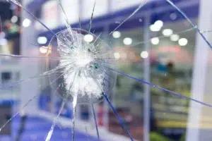 Close-Up Of A Shattered Glass Panel With A Central Impact Point And Radiating Cracks, Against A Blurred Indoor Background.