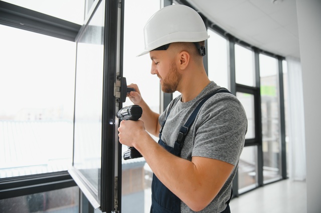 Construction Worker Installing A Window Using A Power Drill, Wearing A Hard Hat And Overalls.