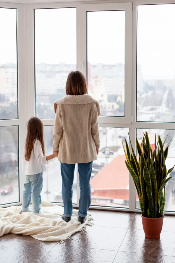 A mother and daughter standing by a large window, looking outside, with a plant on the floor.